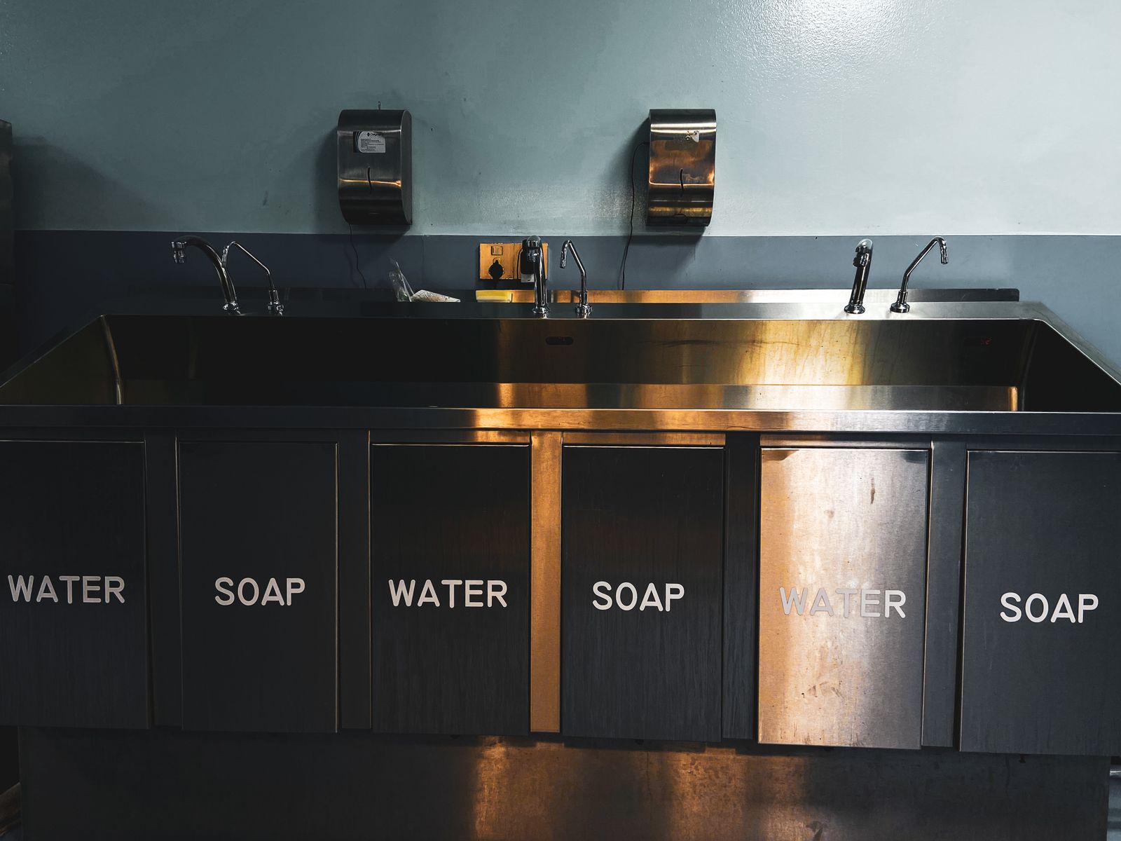Szeroki stalowy zlew z kilkoma kranami i dozownikami do mycia rąk, na frontach napisy water i soap